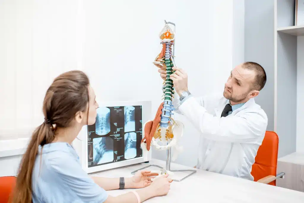 A doctor explaining spine anatomy and surgery options to a patient using a spine model in a consultation room.