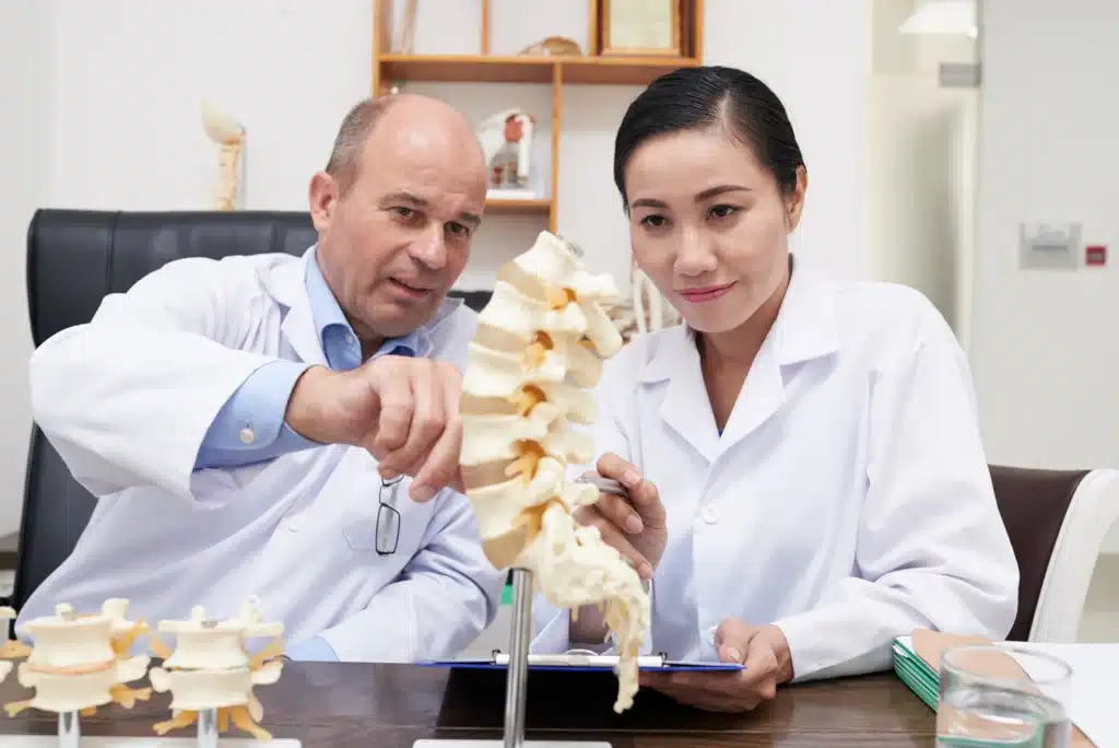 Two doctors analyzing a spine structure model, discussing spinal health and minimally invasive spine surgery options in a clinical setting.