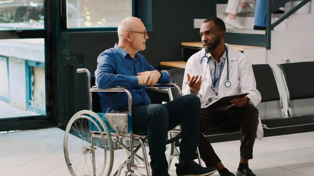 Doctor smiling with senior male patient in a wheelchair after spinal fusion surgery, discussing disability benefits.