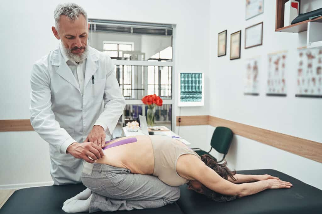 A physiotherapist applies elastic therapeutic tape to a patient's lower back, aiding in muscle recovery after spinal fusion surgery.