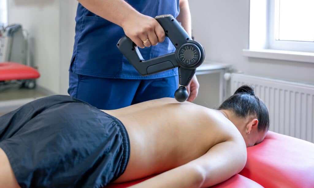 Massage therapy using a percussion massager on a patient's back six months after spinal fusion, helping alleviate discomfort and improve recovery.