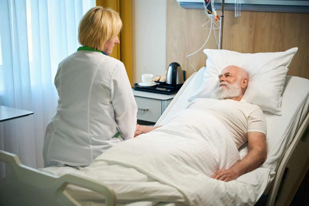 Nurse sitting on the bed beside an elderly patient, providing comfort and post-surgery support.