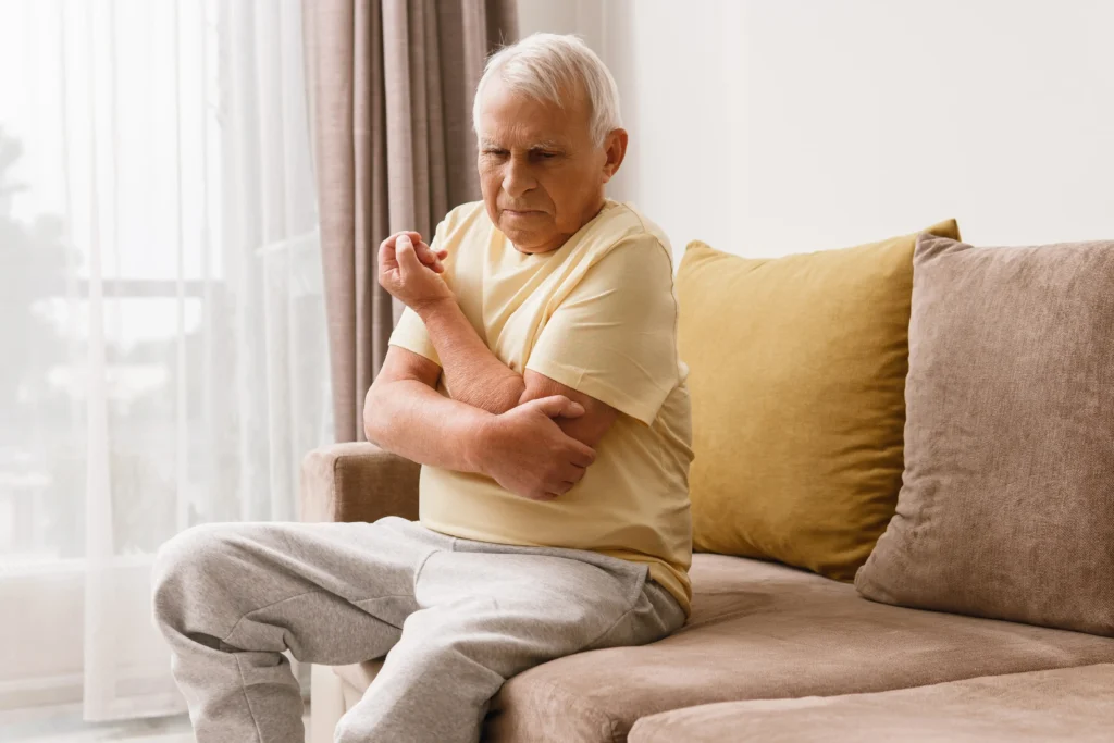 Older man sitting on a couch and experiencing elbow pain, showcasing the importance of geriatric pain management.