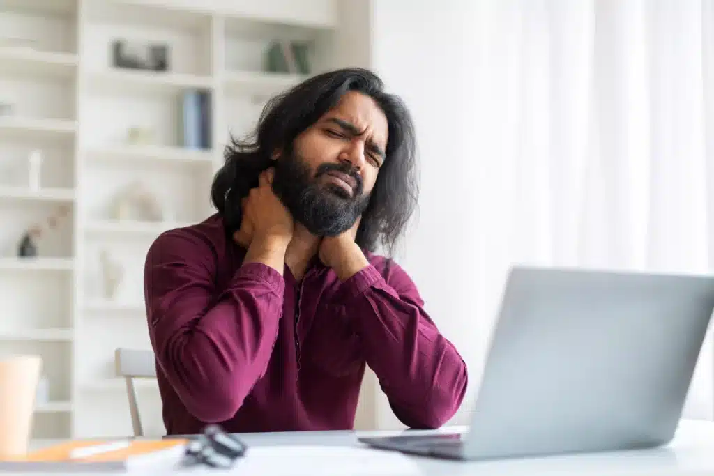 Male freelancer holding his neck in pain while working at a laptop, highlighting the importance of posture and the McKenzie Method.