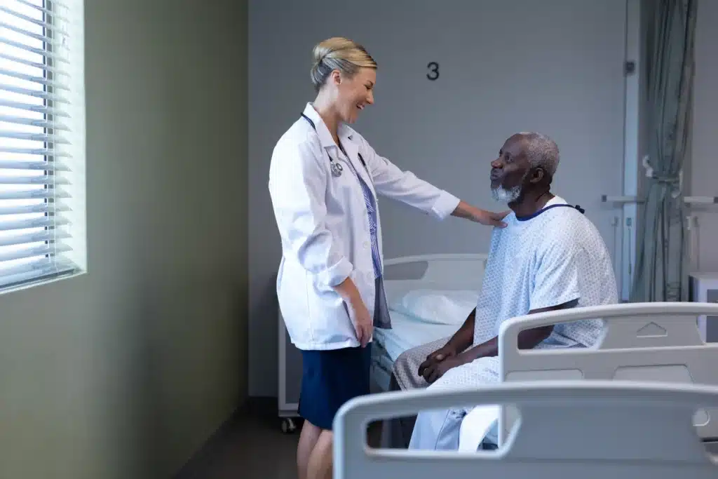 Smiling female doctor in a white coat speaking with an African-American male patient in a hospital room.