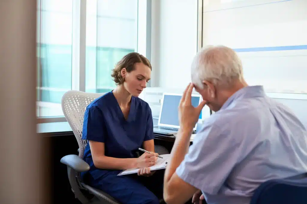 A female healthcare provider writing notes while speaking with a senior male patient experiencing emotional stress.