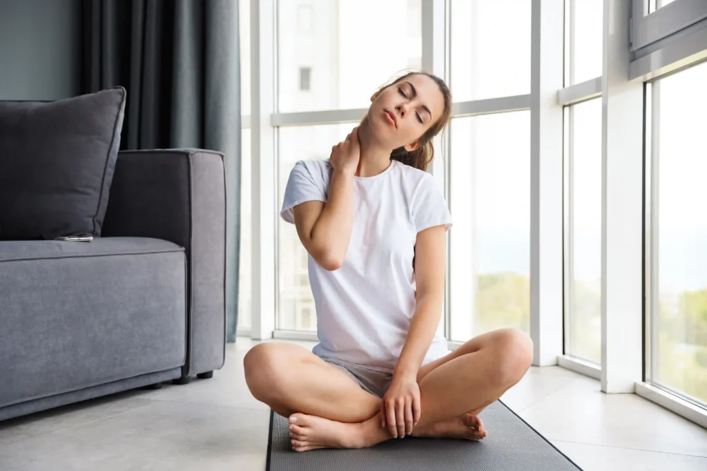 Young woman sitting cross-legged on a yoga mat and stretching her neck for neck pain relief yoga.