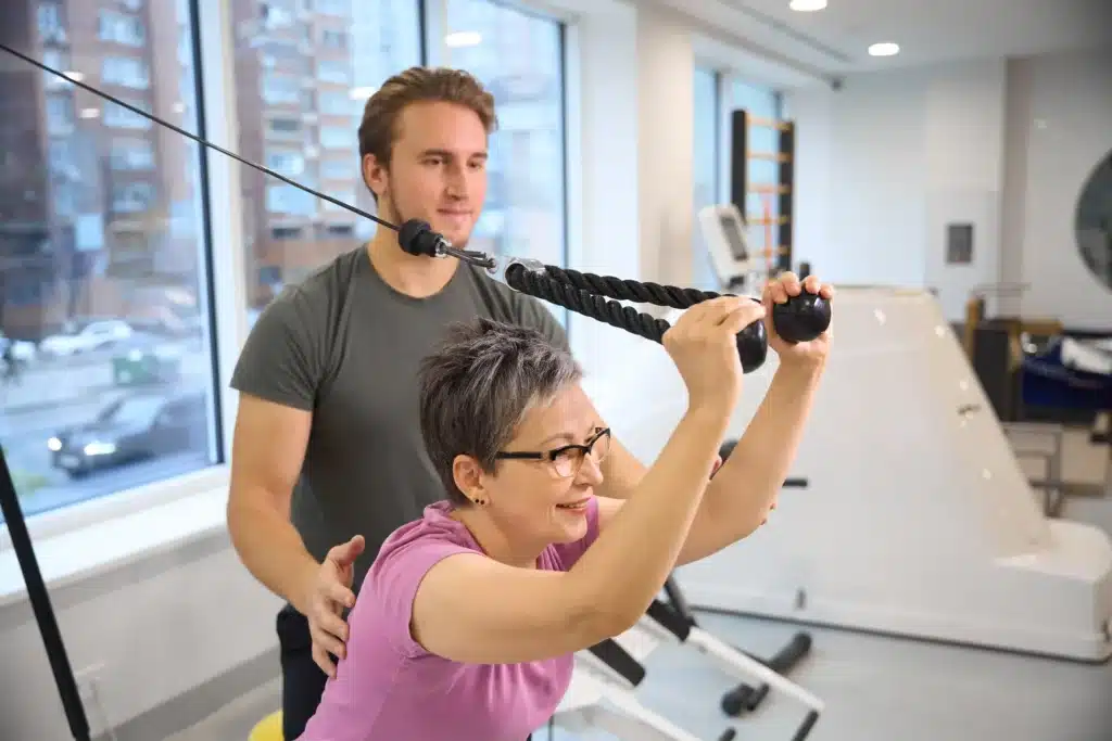 Trainer supporting an adult woman during spinal fusion exercises with a cable machine.