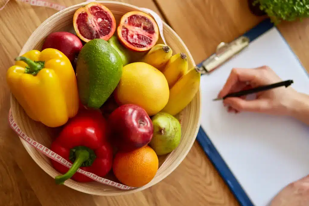 Dietitian writing a personalized nutrition plan with a bowl of fresh fruits and vegetables on the table.