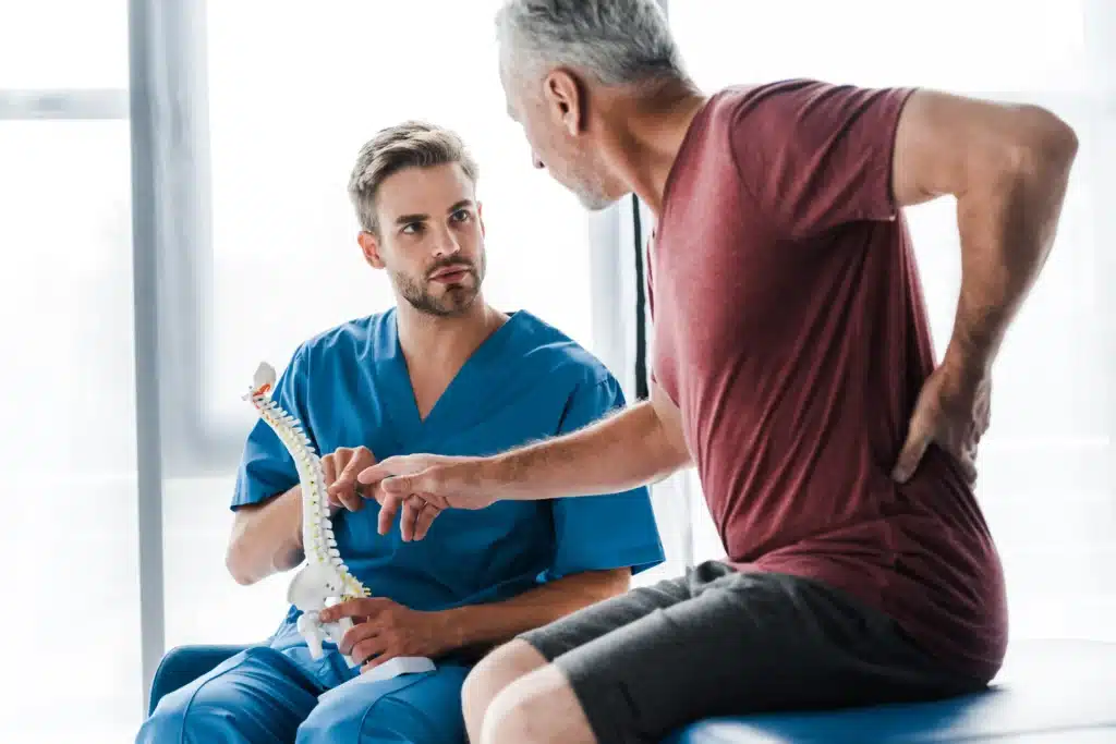 Doctor in blue scrubs showing a spine model to a patient with lower back pain during a consultation.