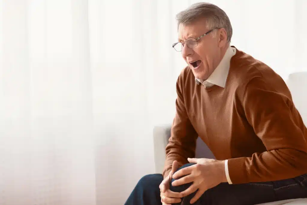 A senior man experiencing knee pain while sitting on a sofa, highlighting the importance of SI joint pain treatment in Long Island.